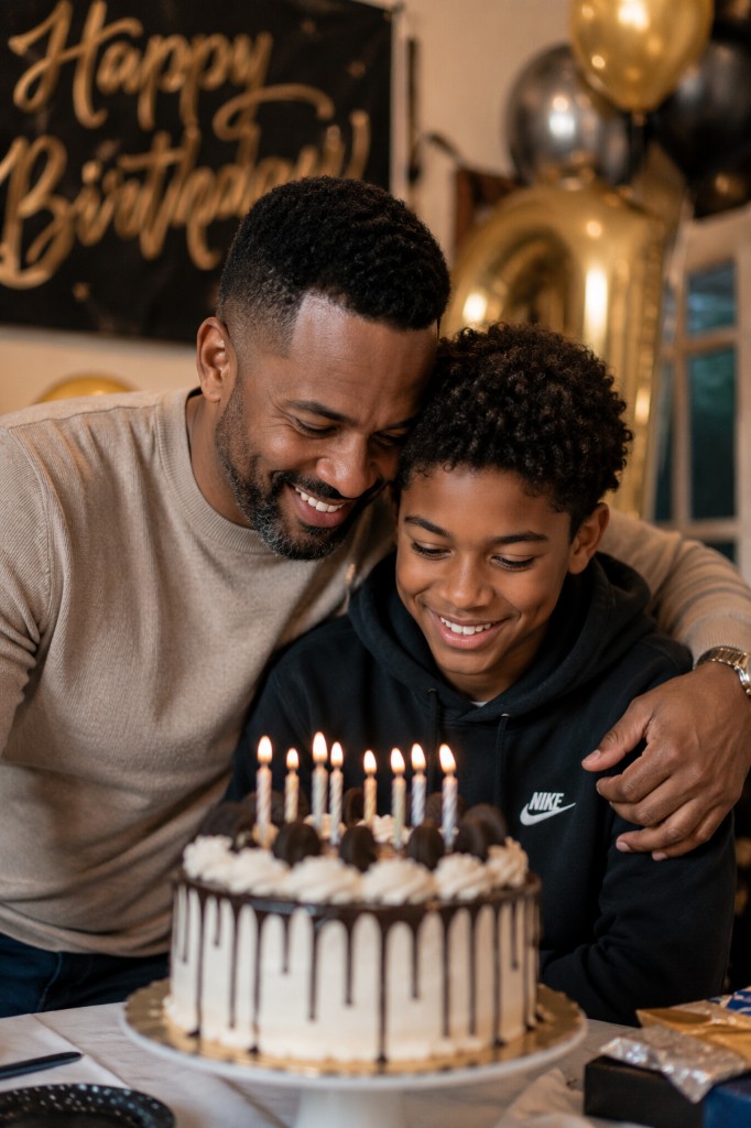 A father and young son smiling together over a lit birthday cake with balloons and a Happy Birthday banner in the background
