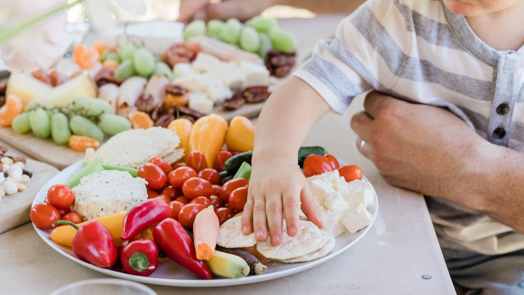 A child's hand reaching toward a colorful platter of crackers, vegetables, and cheese while an adult sits nearby at the table