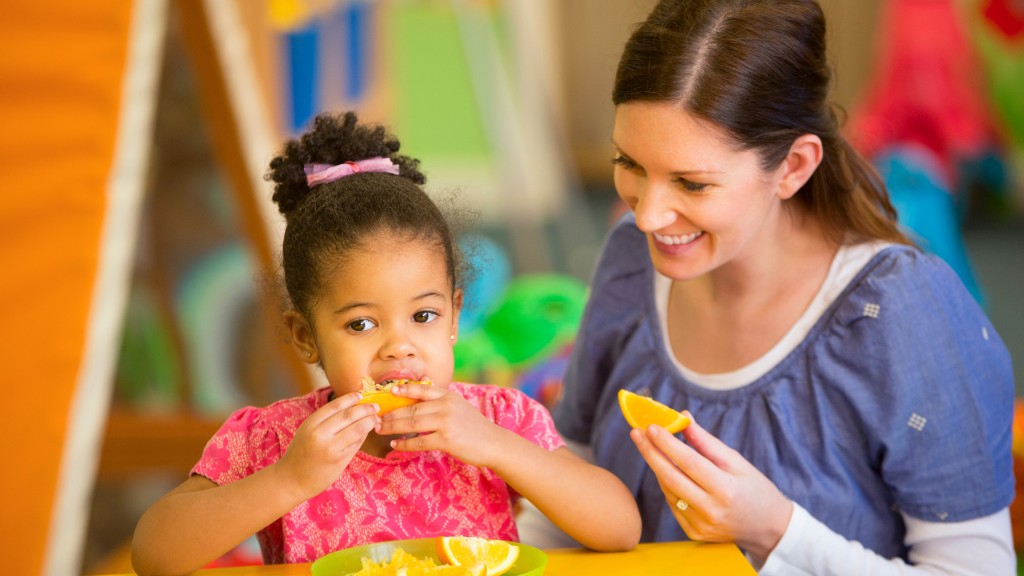A young girl eating an orange slice at a table while a supportive adult smiles and holds another slice beside her in a bright therapy setting