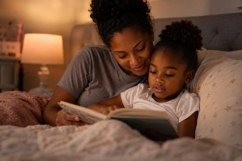 A mother and young daughter reading a book together in bed under warm lamplight