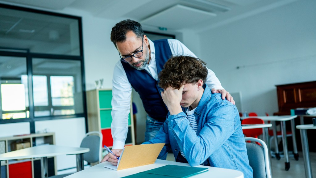 A teacher placing a supportive hand on a student's shoulder while guiding him through work at a classroom desk