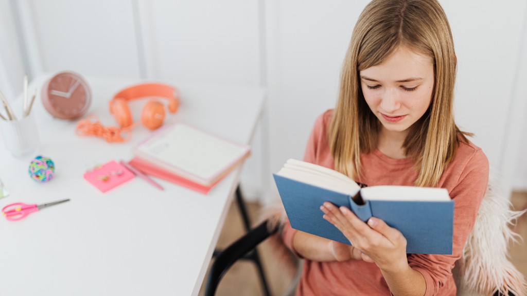 A young girl in a coral shirt sitting at a white desk, focused on reading an open blue book