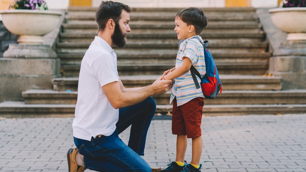 A father kneeling and holding his young son's hands on stone steps as the boy smiles with a backpack on