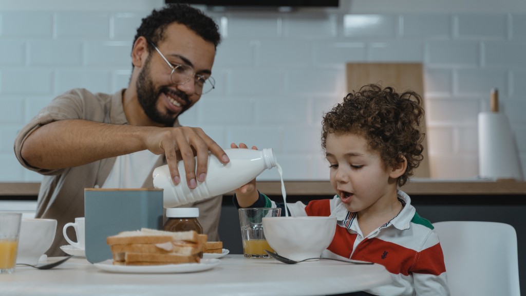 A father pouring milk into a bowl while his young son watches at a breakfast table in the kitchen