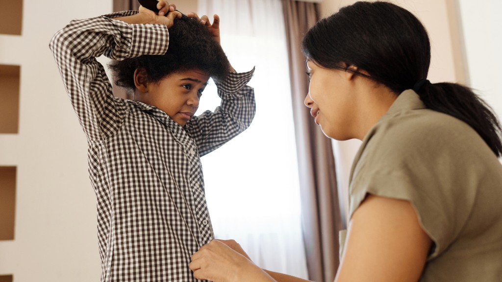 A child concentrating while combing their hair as an adult helps button their checkered shirt