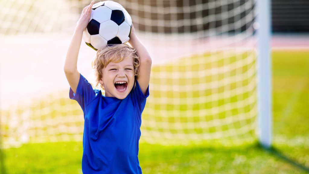 A young boy smiling joyfully while holding a soccer ball above his head on a sunny field