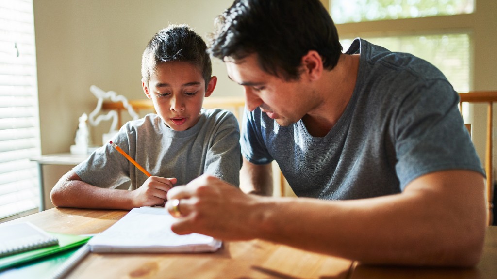 A father pointing at a notebook while helping his son with homework at a wooden table