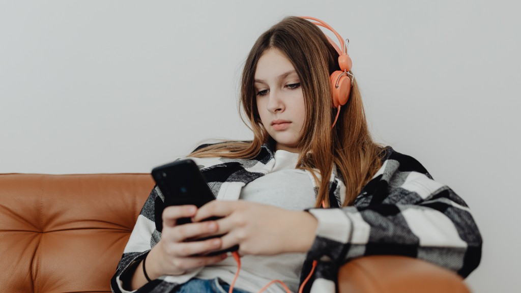 A teenage girl sitting on a couch wearing headphones and looking down at her phone with a pensive expression