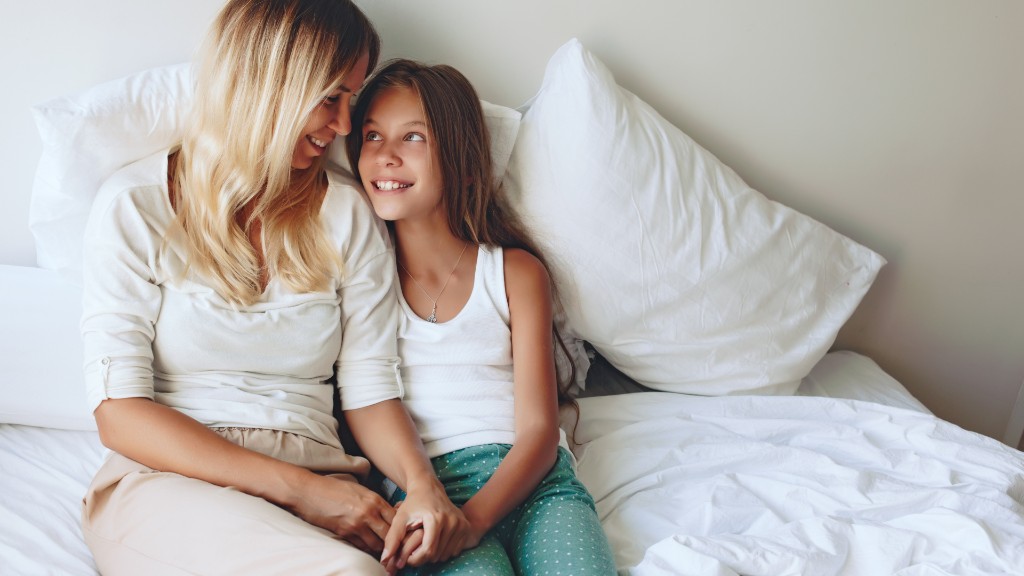 A mother and daughter sitting on a bed holding hands and smiling at each other