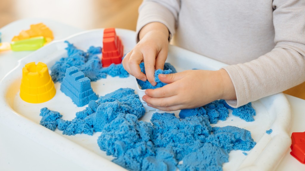 A child's hands playing with blue kinetic sand and colorful molds in a white plastic tray