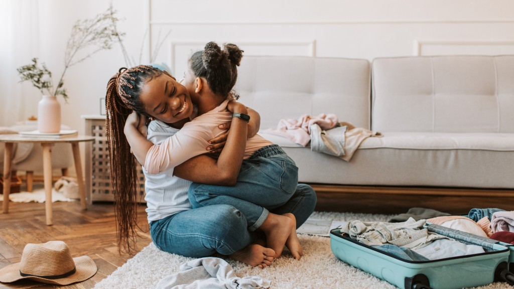 A mother and young daughter kneeling on a rug in a bright living room, embracing in a warm hug