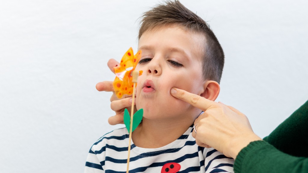 A young boy practicing breath control by blowing a pinwheel during a speech therapy exercise with an adult's hands gently guiding his cheeks