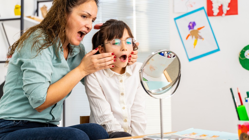 A speech therapist guiding a young girl through mouth-position practice using a tabletop mirror in a classroom setting