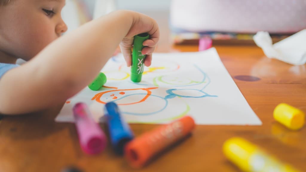 Child drawing with colorful paint sticks on a wooden table